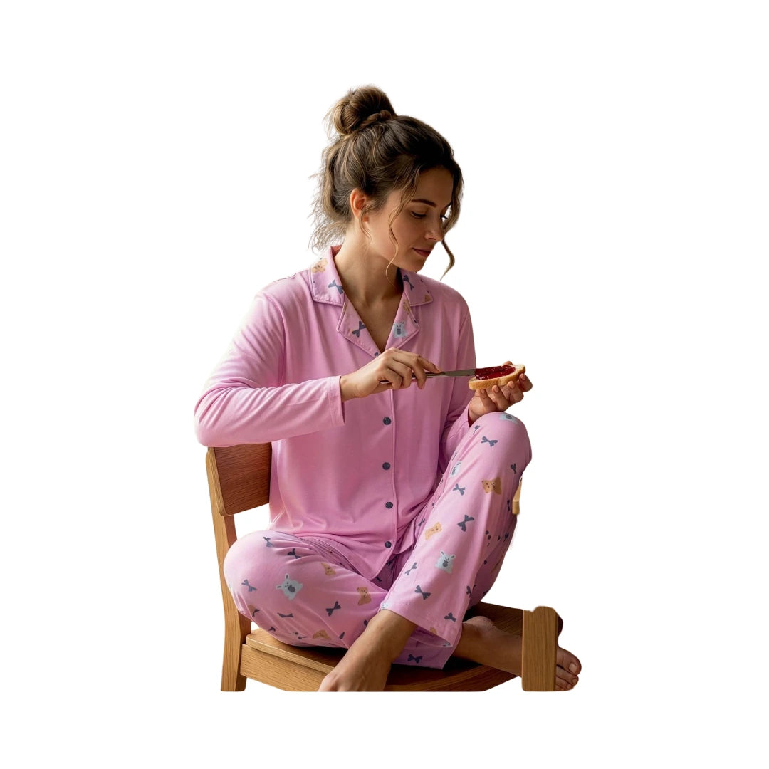 Woman wearing a pink 100% cotton button-up long sleeve pajama set with matching printed full-length pants, seated on a wooden chair against a white background.